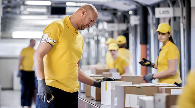 Mann verpackt Kartons in Logistikzentrum, andere Arbeiter im Hintergrund