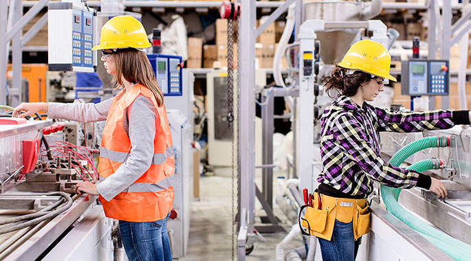 Zwei Frauen in Schutzausr&uuml;stung arbeiten an Maschinen in Produktionshalle