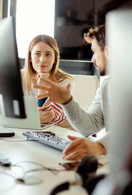 Person erkl&auml;rt gestikulierend am Schreibtisch mit Monitoren, andere h&ouml;rt zu