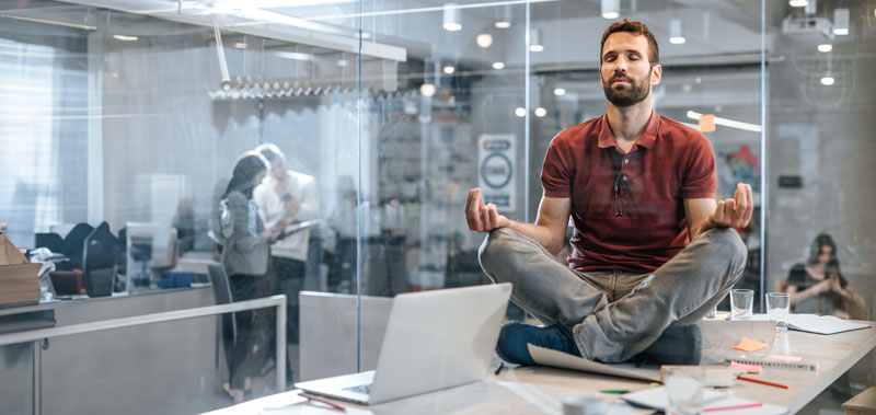 Mann meditiert im B&uuml;ro und sitzt im Schneidersitz auf dem Tisch
