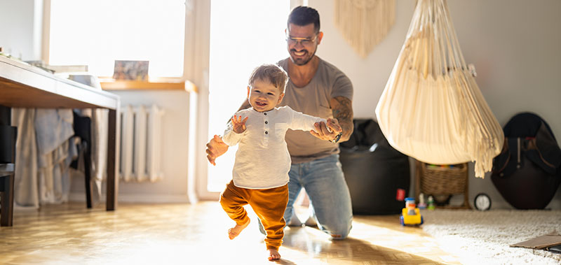 Vater unterst&uuml;tzt sein Kind beim Laufenlernen zu Hause